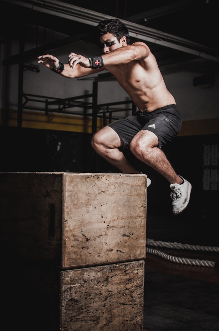 about-img-01 Shirtless man showcasing strength and fitness with a box jump in an indoor gym setting.